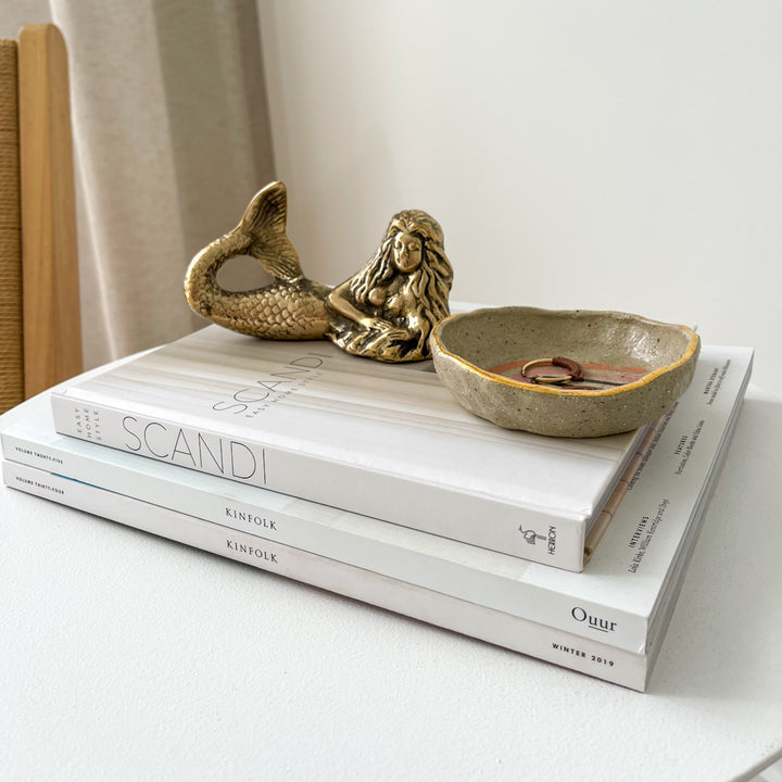A brass mermaid figurine in a lying position, placed on top of a stack of white books, with a decorative bowl beside her.